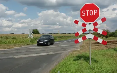 Przejazd kolejowy z zaporami i znakiem STOP. Samochód zbliża się do torów przez pole kukurydzy.