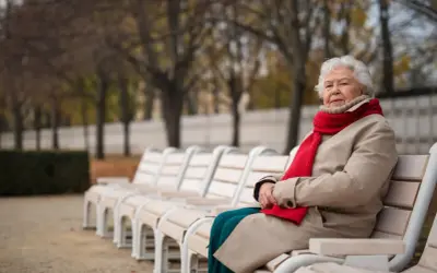 Starsza pani z bystrością umysłu siedzi na ławce w parku, otulona czerwonym szalem i beżowym płaszczem.