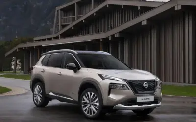 Elegancki, beżowy Nissan X-Trail 2015 zaparkowany przed nowoczesnym budynkiem w górach.