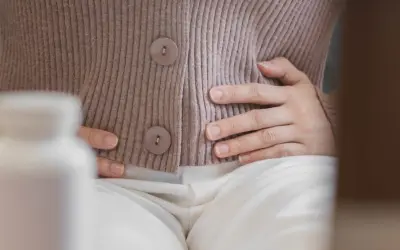 Kobieta trzyma się za brzuch, szukając ulgi na wzdęcia. Obok stoi biała butelka z lekami.