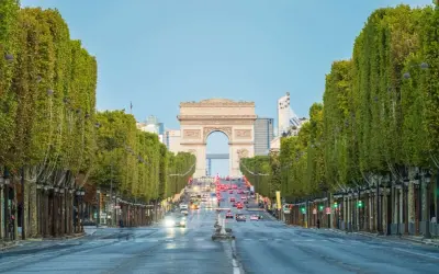 Aleja **Champs-Élysées** w Paryżu z Łukiem Triumfalnym w oddali. Drzewa tworzą zielony tunel, a ruch samochodowy płynie w stronę monumentu.
