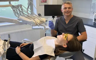 Dentysta w rękawiczce trzyma wiertło, pacjentka w okularach ochronnych leży na fotelu.