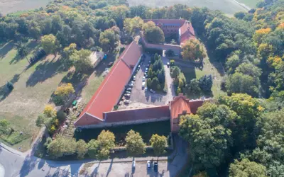 Zamek Bierzgłowski z lotu ptaka, otoczony jesiennym lasem. Widać czerwone dachy, dziedziniec z samochodami i otaczające tereny zielone.