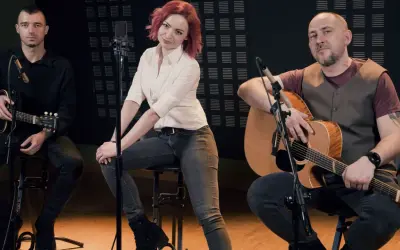 Zespół wykonuje brathanki utwory. Dwóch mężczyzn gra na gitarach, a kobieta z rudymi włosami siedzi pośrodku.