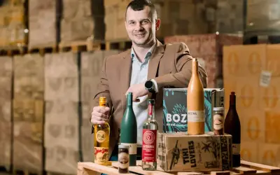 Mężczyzna w brązowej marynarce prezentuje butelki z alkoholem, w tym te z etykietą "Spice Bros". W tle magazyn pełen towarów, sugerujący dystrybucję california alkohol.
