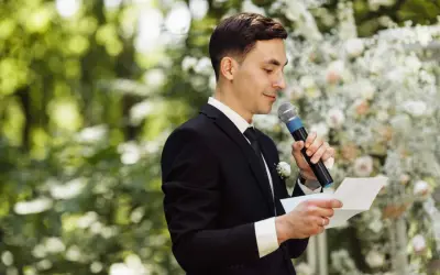 El maestro de ceremonias, con traje negro y micrófono, lee un discurso frente a un arco de flores.