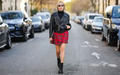 Kobieta w skórzanej ramonesce, czarnym golfie i czerwonej spódnicy w kratę prezentuje styl ubioru grunge. Na nogach ma ciężkie buty.