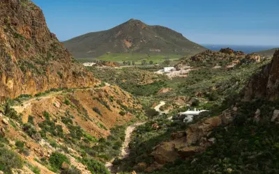 Paisaje árido con el **Cerro Negro** al fondo. Un camino serpentea entre barrancos y vegetación baja, llegando a unas casas blancas.