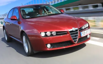 Czerwone Alfa Romeo 159 1.9 JTD pędzi po drodze, kierowca skupiony na trasie.