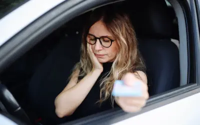 Kobieta w okularach siedzi za kierownicą, trzymając kartę. Wygląda na zmartwioną, jakby czekała na kontrolę, bo grozi jej jazda bez prawa jazdy.