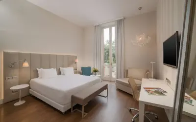 Elegancki pokój hotelowy z dużym łóżkiem, biurkiem i fotelem. Widok na taras z zielenią. Idealne miejsce na wypoczynek w kurorcie na riwierze francuskiej.