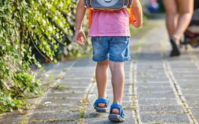 Małe dziecko w różowej koszulce i dżinsowych szortach z plecakiem idzie chodnikiem. To widok, kiedy dzieci idą do szkoły.