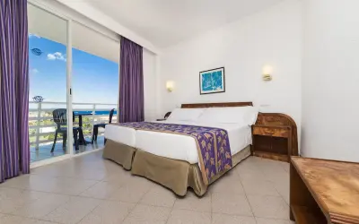 Pokój w hotelu Globales Palmanova Palace z widokiem na morze. Dwa łóżka, fioletowe zasłony i balkon.