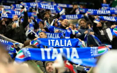 Tłum kibiców na meczu, trzymający niebieskie szaliki z napisem "ITALIA" i flagą Włoch. Emocje towarzyszące kibicowaniu podczas mistrzostw świata.