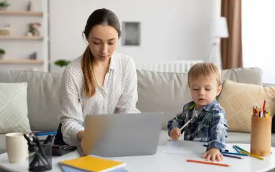 Kobieta pracuje na laptopie, podczas gdy jej syn rysuje obok. To codzienna opieka na dziecko i praca zdalna.