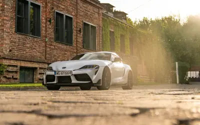 Biała Toyota GR Supra zaparkowana przed ceglanym budynkiem porośniętym bluszczem. Słońce przebija się przez drzewa, tworząc ciepłą, złotą poświatę.