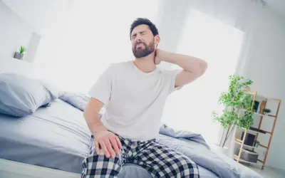 Uomo con torcicollo dopo aver dormito, si massaggia il collo con espressione sofferente, seduto sul letto.