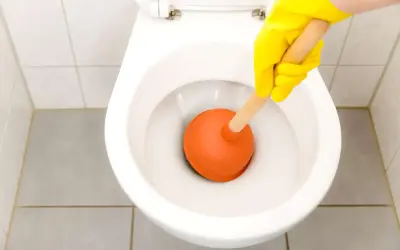 Mano con guanto giallo usa uno stantuffo arancione per liberare un filo d'acqua che scende nel water.