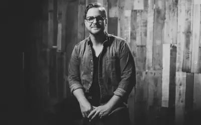 Jordan Reynolds, a talented songwriter, poses confidently in a denim shirt against a rustic wooden backdrop.