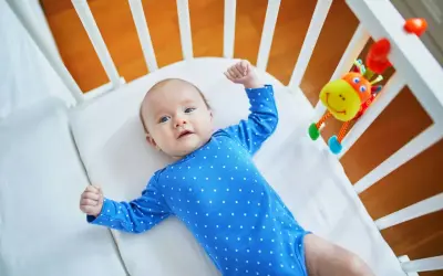 Maluch w niebieskim bodziaku w kropki, leżący w łóżeczku, przeżywa skok rozwojowy 10 tydzień. Z radością podnosi rączki.