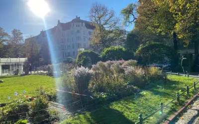 Grüner Park mit Blumenbeeten, Bäumen und einem Altbau unter strahlender Sonne.
