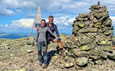 Två glada vandrare vid en stenröse på en bergstopp i Sverige. En modern, pyramidformad byggnad med solpaneler syns i bakgrunden. Perfekt för att utforska Sveriges vandringsleder.