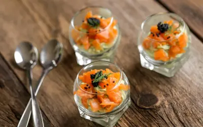 Trois verrines de saumon, avocat et caviar, garnies d'aneth, sur une table en bois. Deux cuillères argentées sont posées à gauche.