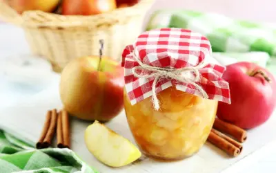 Frisch eingekochtes Apfelkompott im Glas, dekoriert mit kariertem Stoff und Zimtstangen. Äpfel im Korb im Hintergrund.