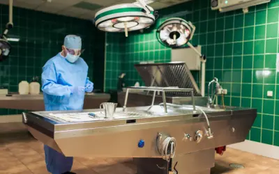 Specjalista w niebieskim fartuchu i masce przygotowuje się do zabiegu tanatopraksji w sterylnym pomieszczeniu z zielonymi kafelkami.