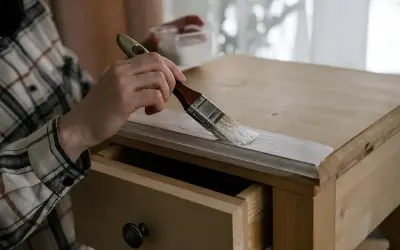 Persona pintando muebles de madera sin lijar, aplicando una capa de pintura blanca con brocha a un mueble de pino.