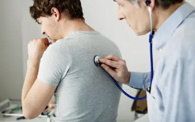 Arzt untersucht Patienten mit Stethoskop. Der Patient hustet, was auf Kurzatmigkeit hindeuten könnte.