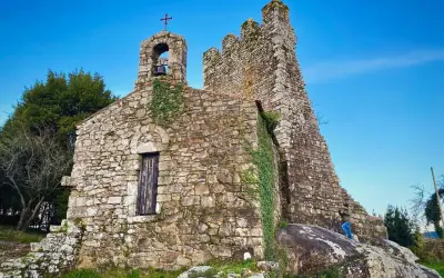 Antigua ermita de piedra con campanario y cruz, rodeada de vegetación. Las **torres de oeste** se alzan imponentes contra el cielo azul.