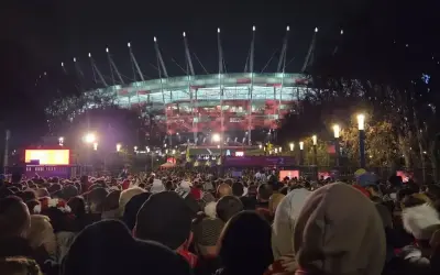 Tłum ludzi zmierza na roztanczony PGE Narodowy. Stadion oświetlony na czerwono, nocna atmosfera.
