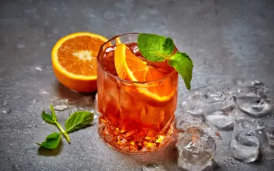 Un cocktail aranciato con fette d'arancia e menta, circondato da cubetti di ghiaccio per cocktail che non si scioglie, su uno sfondo grigio.
