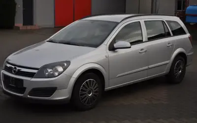 Srebrny Opel Astra H 1.6 benzyna, kombi, zaparkowany na kostce brukowej przed budynkiem z czerwonymi drzwiami.