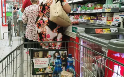 Kolejka w sklepie, ludzie robią zakupy. Widać ceny produktów, przypominając o zakazie handlu w niedziele i święta.