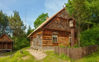 Drewniane chaty i kapliczka na słupie w jednym z **skansenów w Polsce**. W tle las i błękitne niebo.