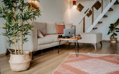 Przytulne, **małe mieszkanie w stylu boho** z beżową kanapą, drewnianym stolikiem i roślinami. Schody prowadzą na piętro.