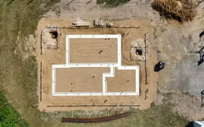 Widok z lotu ptaka na budowę domu. Widać gotowy fundament, piasek, wykopy i materiały budowlane.