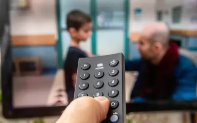 Dłoń trzymająca pilot do telewizora, na ekranie widać scenę z dzieckiem i mężczyzną. Czas na ulubiony serial, opłacony abonament radiowo-telewizyjny działa!