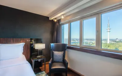Pokój w Blu Radisson Hamburg z widokiem na wieżę telewizyjną. Wygodne łóżko, fotel i stolik nocny.