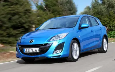 Niebieska Mazda 3 2010 w wersji hatchback pędzi po krętej drodze wśród zieleni.
