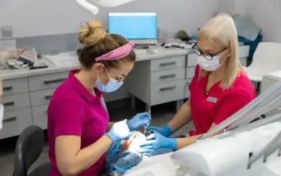 Dwie dentystki w maseczkach i rękawiczkach pracują przy pacjencie. Jedna z nich używa narzędzi, druga asystuje. Zabieg bez bólu dzięki lidokainie.