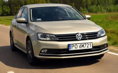 Złoty VW Jetta MK6 jedzie po drodze wśród zielonych pól i lasów.