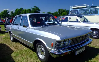 Srebrny fiat 130 z lat 70. prezentuje się elegancko na trawie, obok innych klasyków.