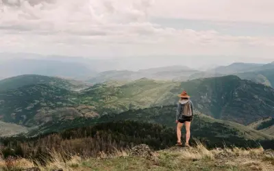Una excursionista con sombrero admira las majestuosas montañas de Madrid, un paisaje verde y brumoso bajo un cielo nublado.