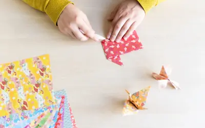Hände falten Papier für eine Origami Kugel Anleitung. Mehrere bunte Origami-Papierbögen liegen bereit.