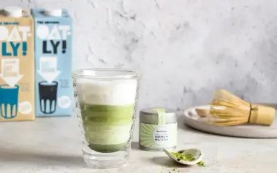 Warstwowy napój matcha z mlekiem owsianym, obok puszka z matchą i bambusowa miotełka. Idealny matcha przepis.