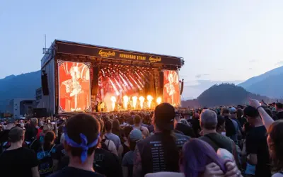 A massive crowd enjoys a performance at the Greenfield festival, with a large screen displaying a performer and flames erupting on stage.