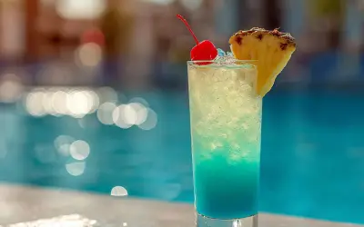 En blå och gul drink med körsbär och ananas står vid en pool. Perfekt för en solig dag.